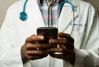 Medical professional in white lab coat and stethoscope using a smartphone; viewed from chest down.
