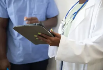 Doctor in white lab coat holding a tablet consults with a colleague in blue scrubs in a clinical setting.