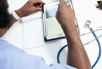 Medical professional in scrubs reviewing patient records on a tablet beside a stethoscope and notepad at a desk.