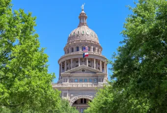 Texas State Capitol dome in Austin framed by green trees; with US and Texas flags flying under a clear blue sky.
