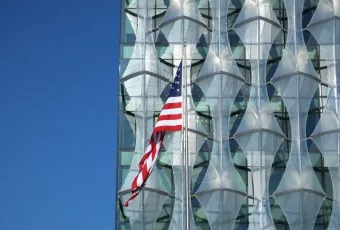 American flag flying in front of the geometric glass facade of the US Embassy in London under a clear blue sky.