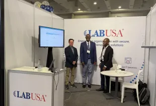 Three LABUSA representatives standing at booth 130 during a trade show; with branded banner and monitor display.