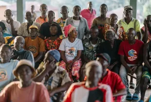 A large group of men and women of various ages seated and standing together at an outdoor community meeting.
