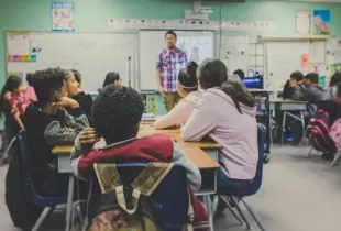 Diverse middle school students seated at desks face a male teacher presenting a lesson on a smartboard.