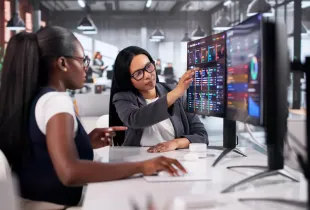 Two professional women reviewing colorful data dashboards on large monitors in a modern office environment.