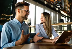 Two business professionals discuss IT solutions in a modern office, one holding a tablet during a collaborative meeting.