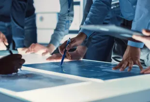 Team of professionals collaborating over a glowing digital display table; reviewing technical diagrams in an office setting.