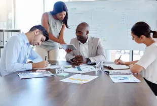 Four professionals collaborate at a conference table with charts and a tablet, a business analysis whiteboard behind them.