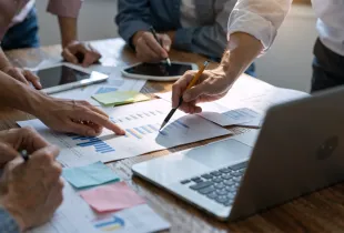 Business professionals reviewing charts; graphs; and data reports on a wooden conference table with laptops and tablets.