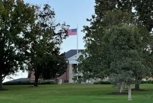 American flag flying above a brick government building with white columns; surrounded by mature trees and manicured lawn.