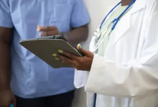 Doctor in white lab coat holding a tablet consults with a colleague in blue scrubs in a clinical setting.