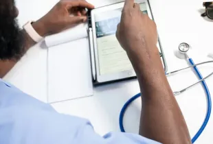 Medical professional in scrubs reviewing patient records on a tablet beside a stethoscope and notepad at a desk.