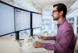 IT professional in purple shirt reviews Quality Management compliance dashboard across multiple monitors in modern office.
