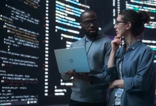 Two IT professionals review source code on a laptop in front of large screens displaying code in a dark room.