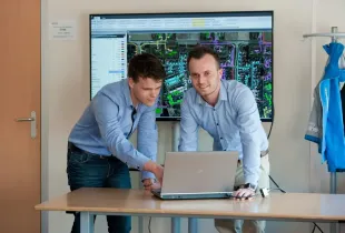 Two professionals in button-down shirts review GIS mapping software on a laptop, with a large network map on screen.