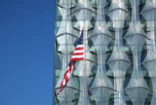American flag flying in front of the geometric glass facade of the US Embassy in London under a clear blue sky.