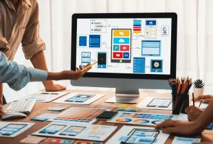 Designers gather around a monitor showing app wireframes, with printed mockups and color swatches spread across the desk.