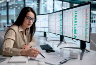 Woman in glasses reviews data dashboards on dual monitors at an enterprise office desk; taking notes with a pen.