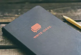 A closed notebook lettered "Write ideas" lying on a wooden desk beside a pencil.