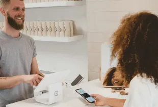 A shopkeeper stands at a small retail counter as a customer pays using a phone.
