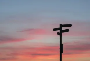 A fingerpost with arms pointing in several directions, silhouetted against a pink sunset sky.