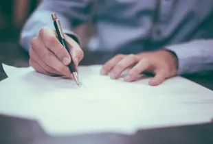 A person leaning over a desk, pen in hand, signing a sheet of paper.