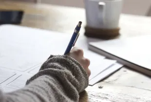 A person writing on a printed document at a wooden desk beside a cup of coffee.