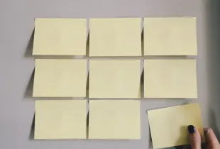 A hand placing a paper note into an ordered grid of notes on a wall.