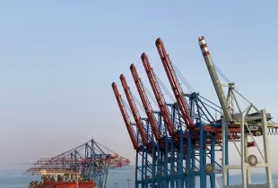 A row of red container cranes standing along a port quay at dusk.