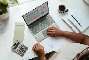 An overhead view of a desk with a laptop showing a budget spreadsheet, a calculator, and a notebook.
