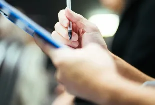 A hand holding a pen over a clipboard while taking notes during an assessment.