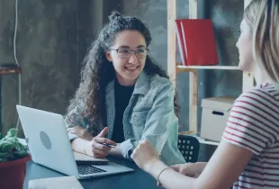 Two colleagues talking over an open laptop at a table in a small workspace.