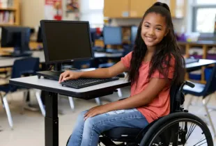 Smiling middle school student in a wheelchair uses a desktop computer at an accessible desk in a K-12 classroom.