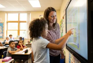 A teacher guides a young student using a touchscreen interactive whiteboard while classmates watch from their desks.