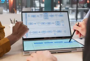 Two professionals review a multi-lane organizational workflow diagram on a dual-screen laptop; with a customer journey map on the lower display.