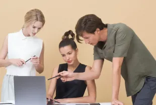 Three colleagues gathered around a laptop, one pointing at the screen while explaining.