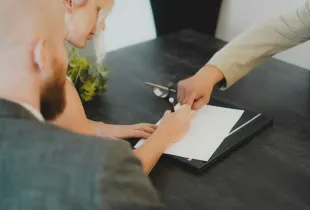 A person signing a document on a folder while two colleagues look on across a meeting table.