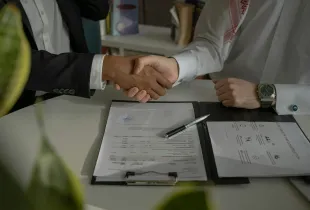 Two people shaking hands over a signed document folder and pen on a desk.