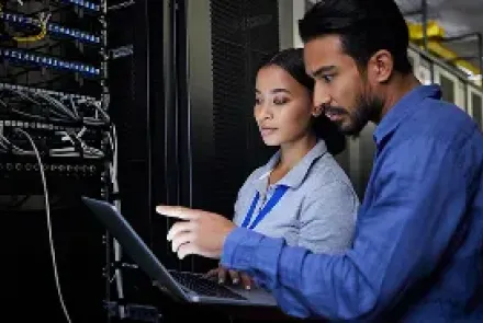 Two IT professionals working on a laptop in a server room