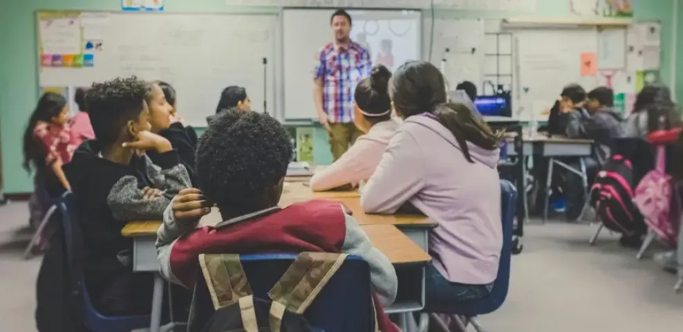 Diverse middle school students seated at desks face a male teacher presenting a lesson on a smartboard.