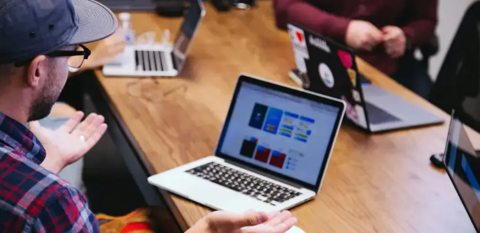 IT professionals collaborate around a wooden table with several laptops open during a team strategy meeting.