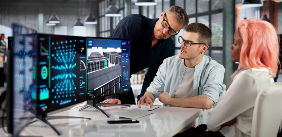 IT professionals collaborate at a workstation reviewing 3D data center and network analytics dashboards on dual monitors.
