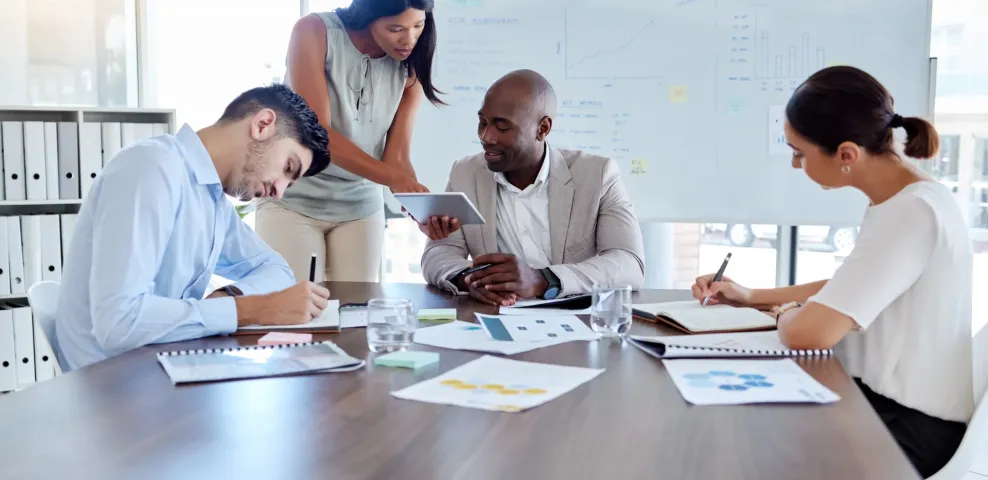 Four professionals collaborate at a conference table with charts and a tablet, a business analysis whiteboard behind them.
