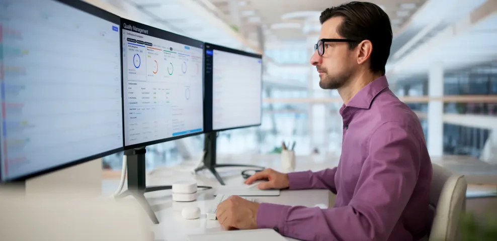 IT professional in purple shirt reviews Quality Management compliance dashboard across multiple monitors in modern office.