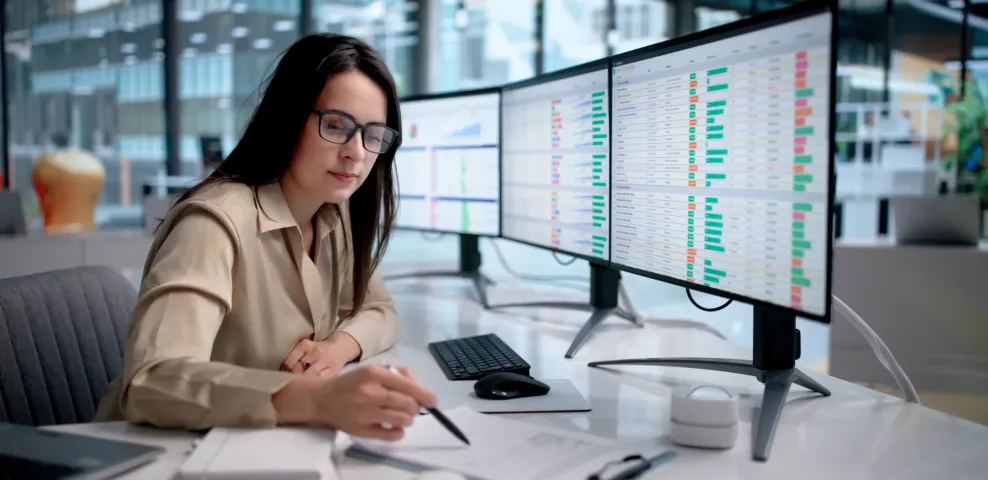 Woman in glasses reviews data dashboards on dual monitors at an enterprise office desk; taking notes with a pen.