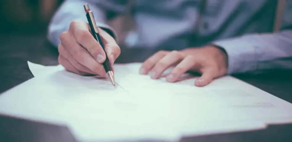 A person leaning over a desk, pen in hand, signing a sheet of paper.