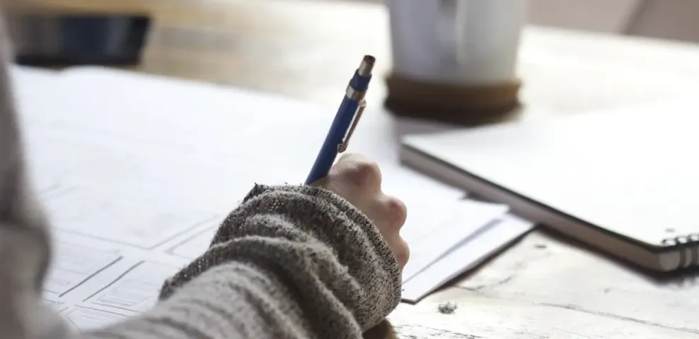 A person writing on a printed document at a wooden desk beside a cup of coffee.