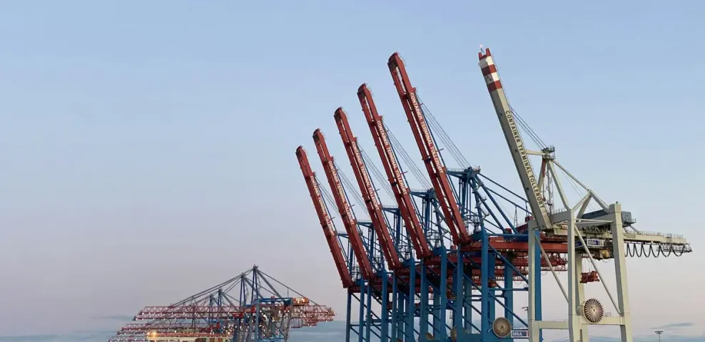 A row of red container cranes standing along a port quay at dusk.