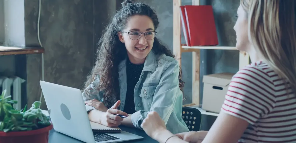 Two colleagues talking over an open laptop at a table in a small workspace.