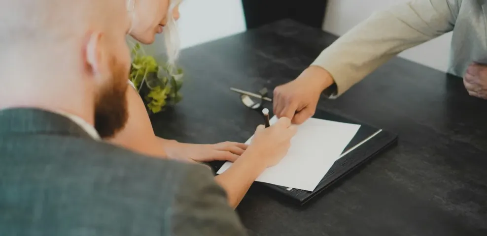 A person signing a document on a folder while two colleagues look on across a meeting table.